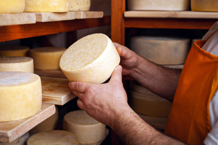 Manos de una persona sostienen un queso; al frente, repisa con más quesos